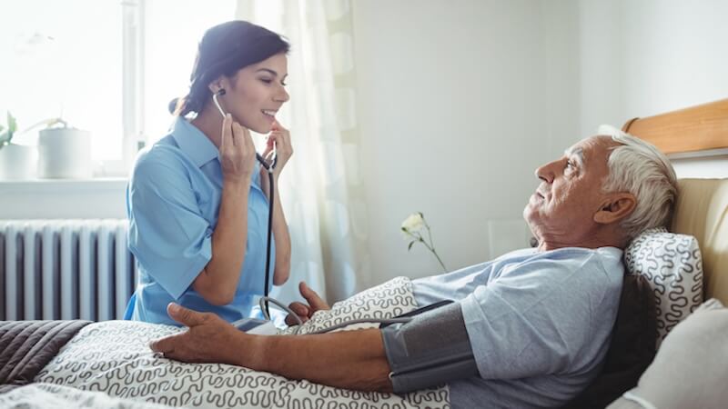 A nurse with a stethoscope checking the blood pressure of an elderly man lying in bed.