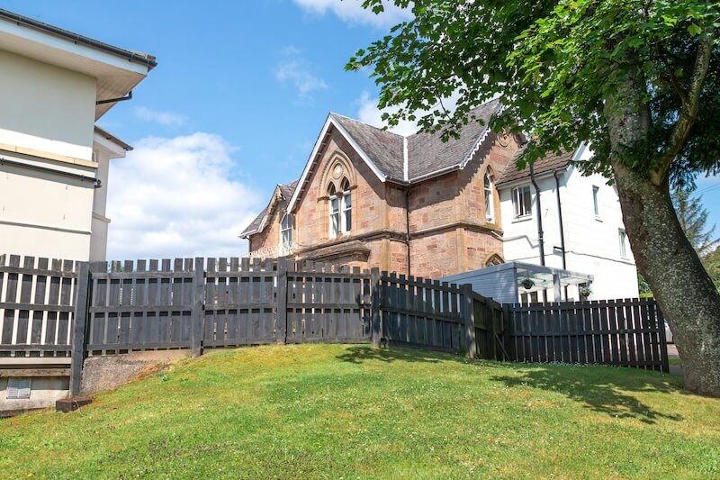 Vibrant green grass in the foreground, a tall tree beside a gray wooden fence, with a care home visible behind the fence.